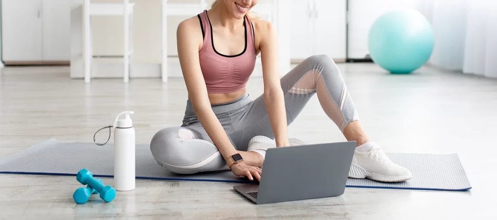 A woman in workout clothes sits on a yoga mat while typing on a laptop in front of her with weights and a water bottle nearby.