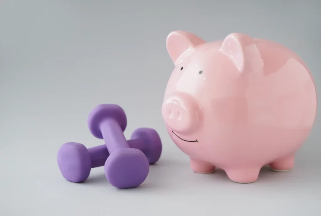 pink piggy bank sitting next to a pair of purple dumbbells on a light grey background, symbolizing fitness savings or financial health in fitness.