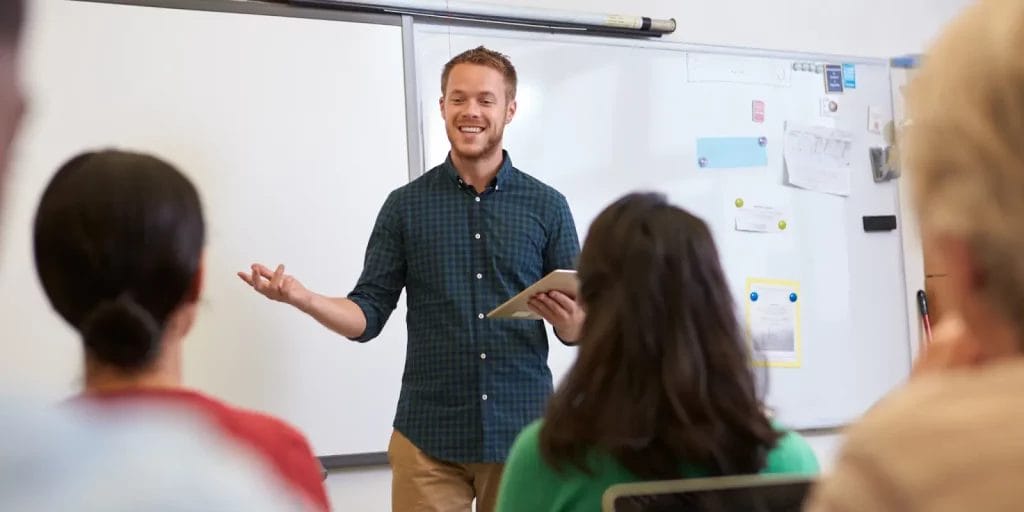 A young man stands at the front of the class, smiling as he teaches, engaging his students.