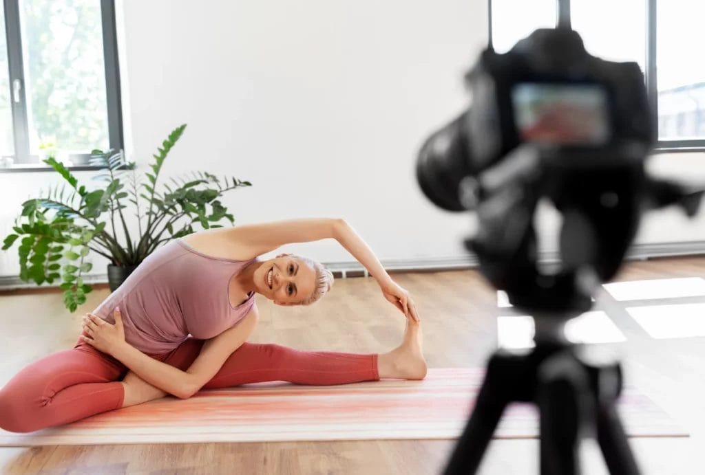 A yoga instructor in pink athleticwear films herself in a seated side bend pose in an open room with a potted plant.