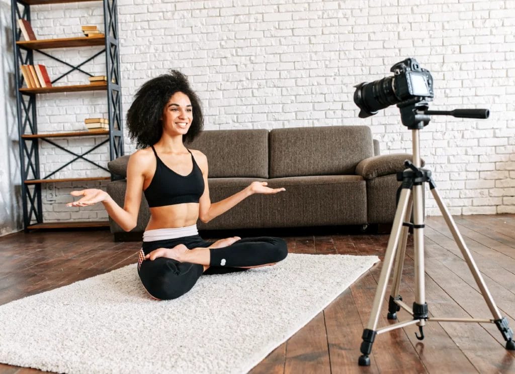 A yoga instructor in black leggings and a black sports bra sits in full lotus pose while facing and smiling at a camera.