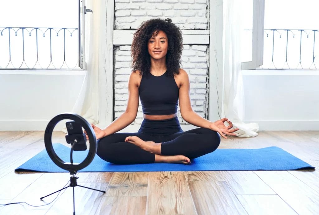 A yogi wearing black leggings and a black crop tank films herself in lotus position with a smartphone and a ring light in front of a white background.