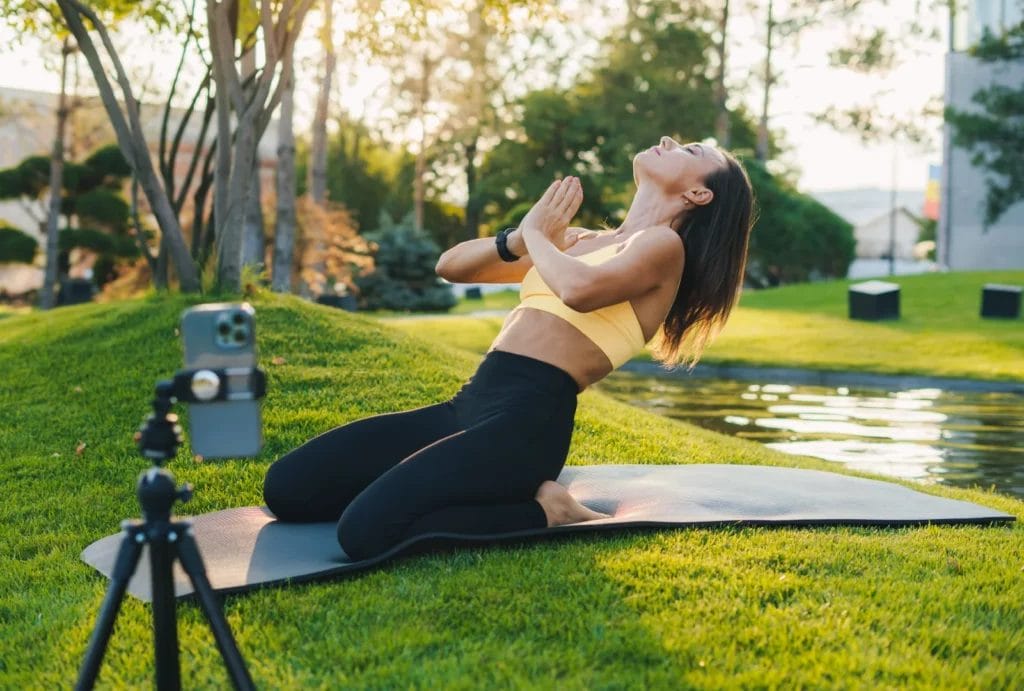Woman recording a kneeling yoga pose outdoors on the grass in front of a pond.
