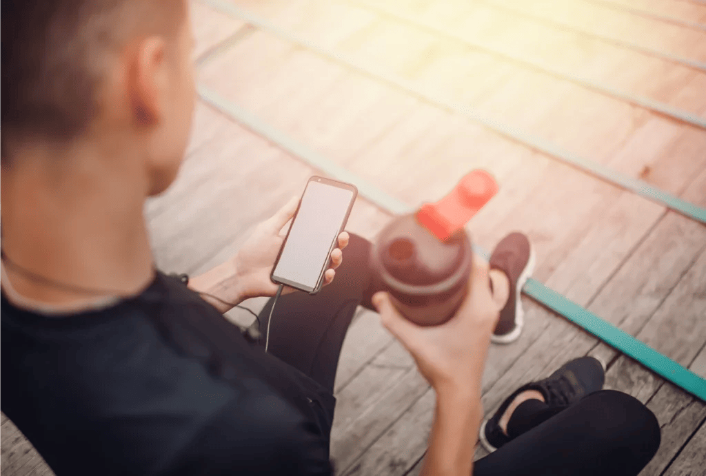 An over-the-shoulder view of a personal training checking his smartphone and holding a shake bottle while sitting down.