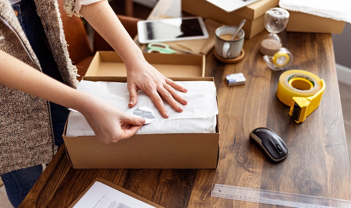 A woman is packaging an online sale by hand on a wooden desk in a work studio in her home.