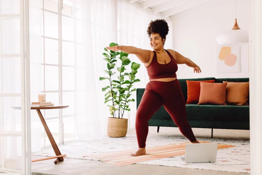 A plus-sized woman confidently practicing warrior pose in a bright, modern living room with a green couch and light décor.