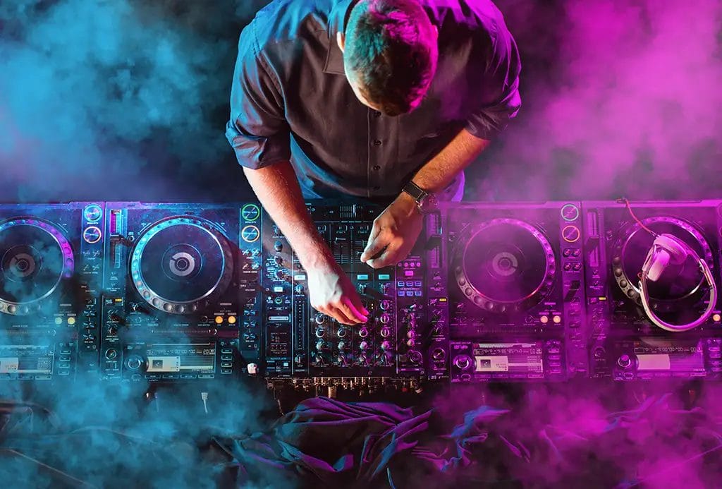 An overhead shot of a DJ at a turntable lit by purple and blue lights and surrounded by fog.