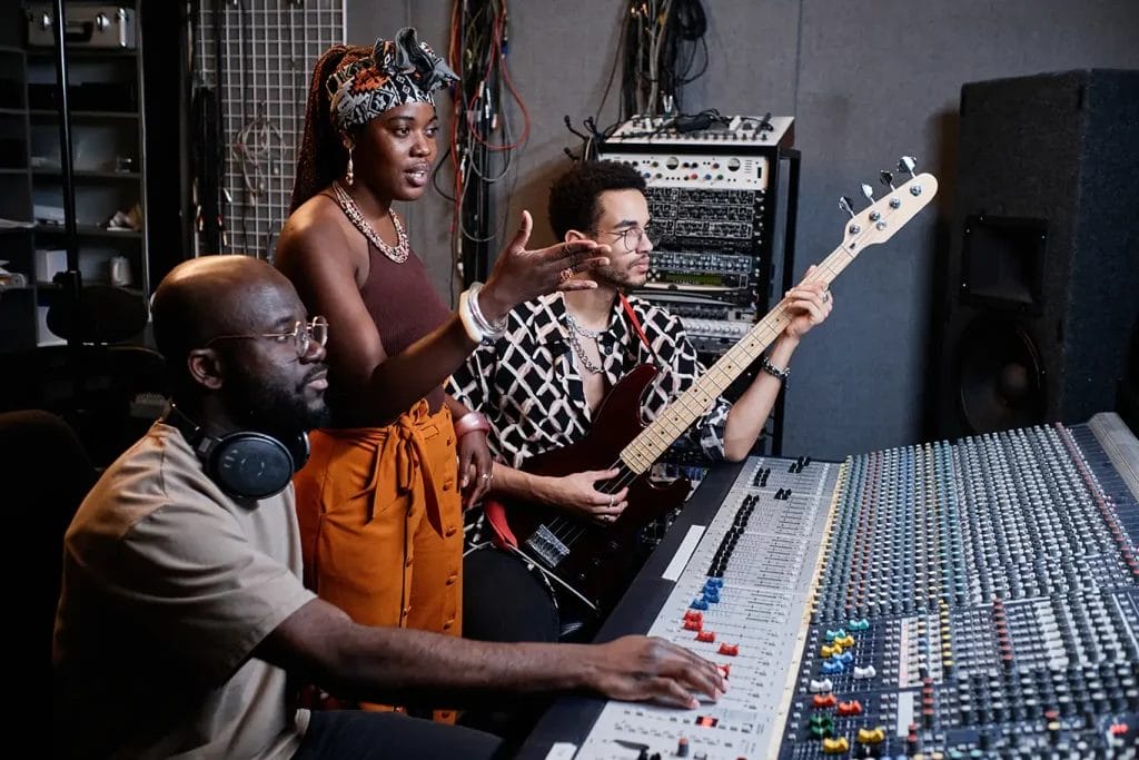 A trio of musicians in a recording studio in front of a soundboard, featuring one playing a guitar and one in the middle speaking, with gray walls.