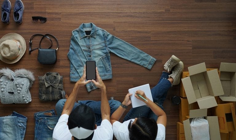 A man and woman sit on the hardwood floors of their home as they photograph different products they are listing on their eBay store.