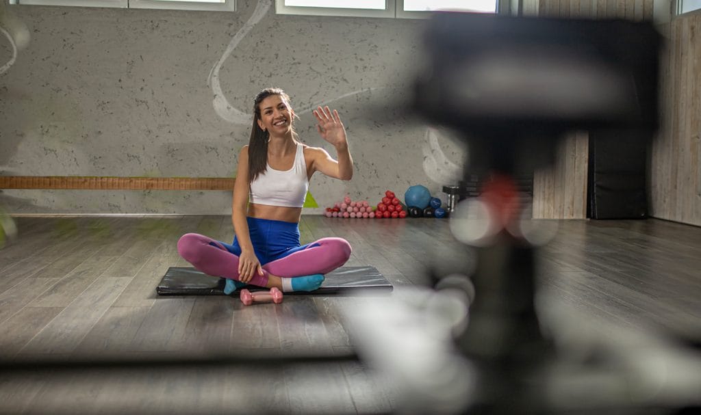 A young woman waves to her camera from her private studio where she films an online personal training video.