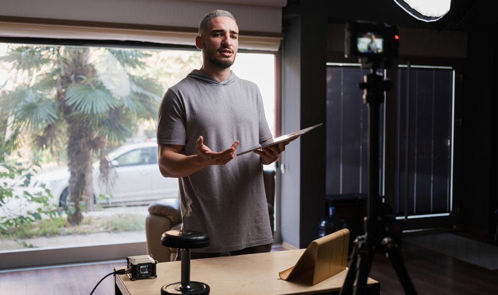 A virtual trainer holds a clipboards and talks to his camera as he goes over some fitness plans for a virtual client.