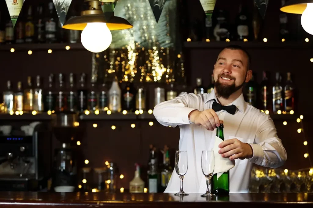 A bartender behind a bar smiles while opening a champagne bottle to serve to customers.