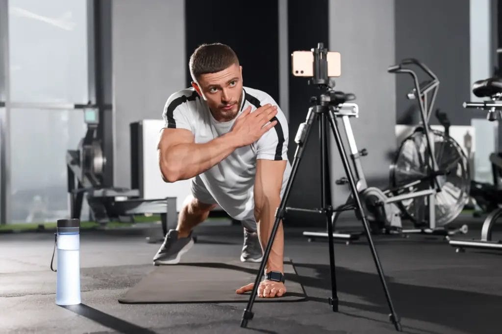 A personal trainer wearing a white-and-black t-shirt and gray sneakers performs elbow taps in plank while filming content using a smartphone and tripod in a fitness studio.
