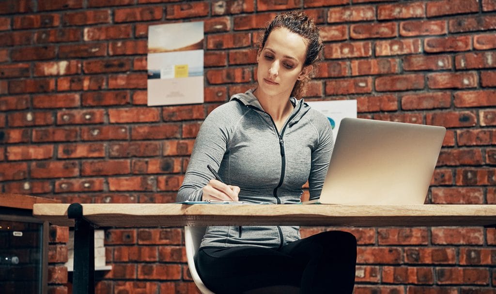 A young woman sits at a desk in a fitness room while she works on her personal trainer certification program from her laptop.