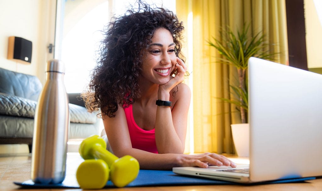 A young woman lays on a yoga mat in her home and is propped up on her elbows as she uses one hand to scroll through the curriculum of her personal trainer certification course.