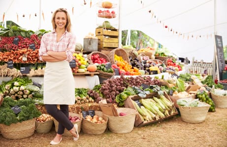 A farmers' market vendor is proudly standing in front of her booth that is fully stocked and decorated for an event.