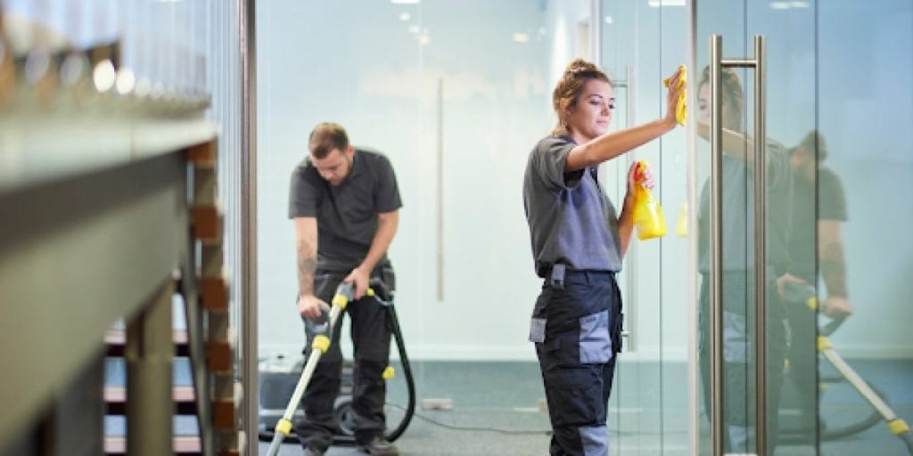 a woman cleaning a glass door in an office building while a man vacuums the floor behind her