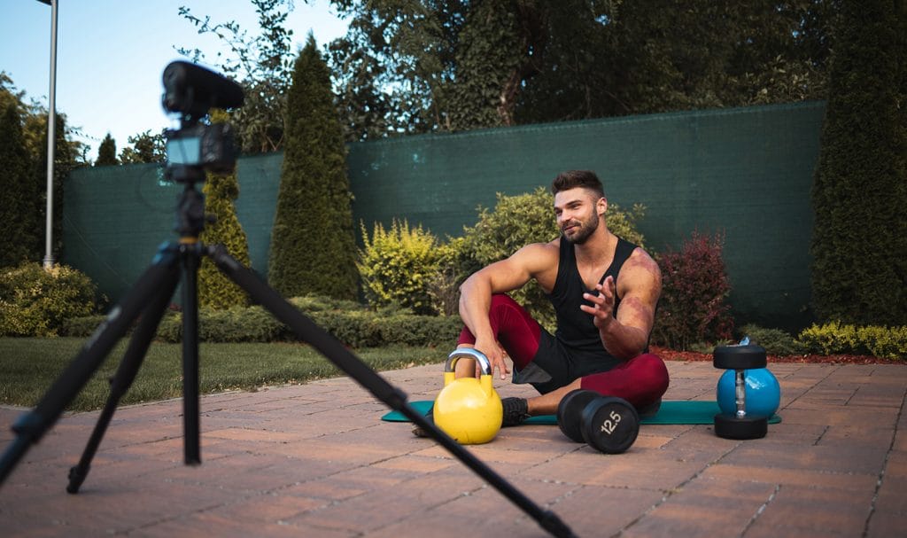 A young man sits on a mat in his backyard with kettlebells and weights as he speaks toward his camera on a tripod for an online personal training video recording.