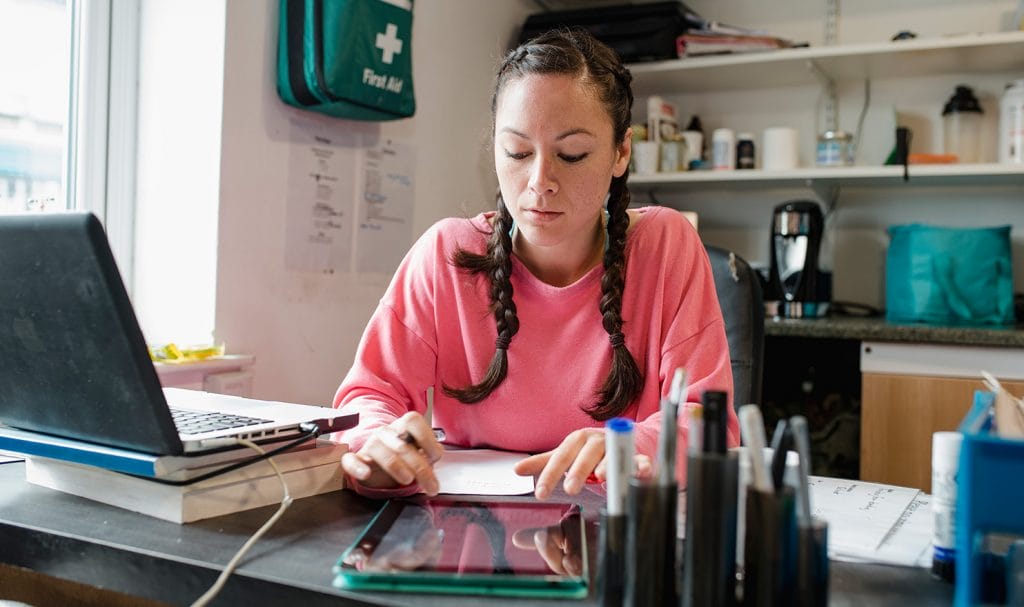 A virtual personal trainer sits at a desk in her office with a laptop and notepad beside her as she works on creating online content on a tablet.