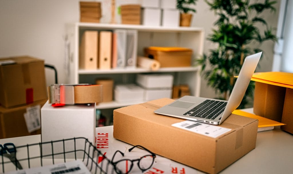 An open laptop sits on a package on a desk in a home office where a small business owner operates from. There are glasses and office supplies on the desk with more packages awaiting shipment on a shelf behind the desk.