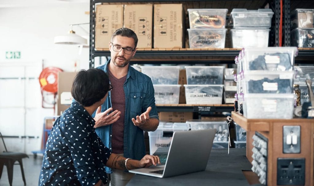 Two small business employees are discussing a new project together and organizing their tasks together. They are in a workspace with lots of business supplies and inventory on desks and shelves around them.