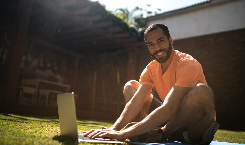 A virtual personal trainer smiles as he sits in his backyard on his laptop where he is organizing his client workout plans.