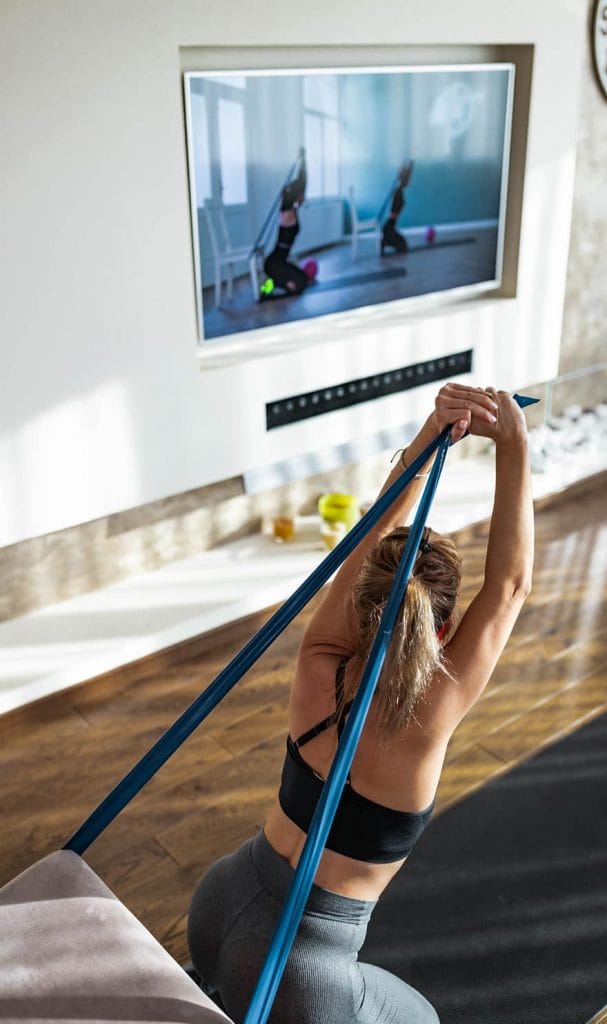 An online personal trainer client is using a resistance band on her chair to exercise along to an online training video.