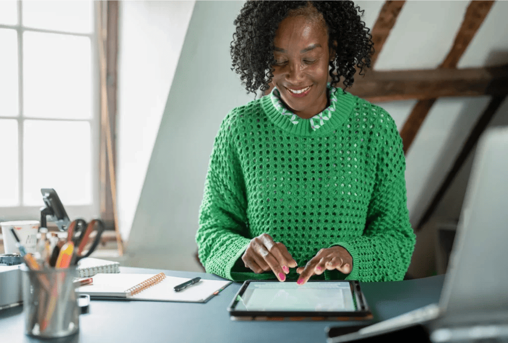 A woman in a green sweater smiles while using a tablet in a well-lit workspace, with stationery and a notebook placed on the desk.