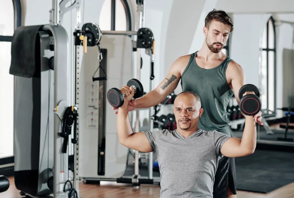 A personal trainer helps a client with lifting dumbbells during a seated shoulder press workout in a gym setting. The trainer watches closely to ensure proper form.