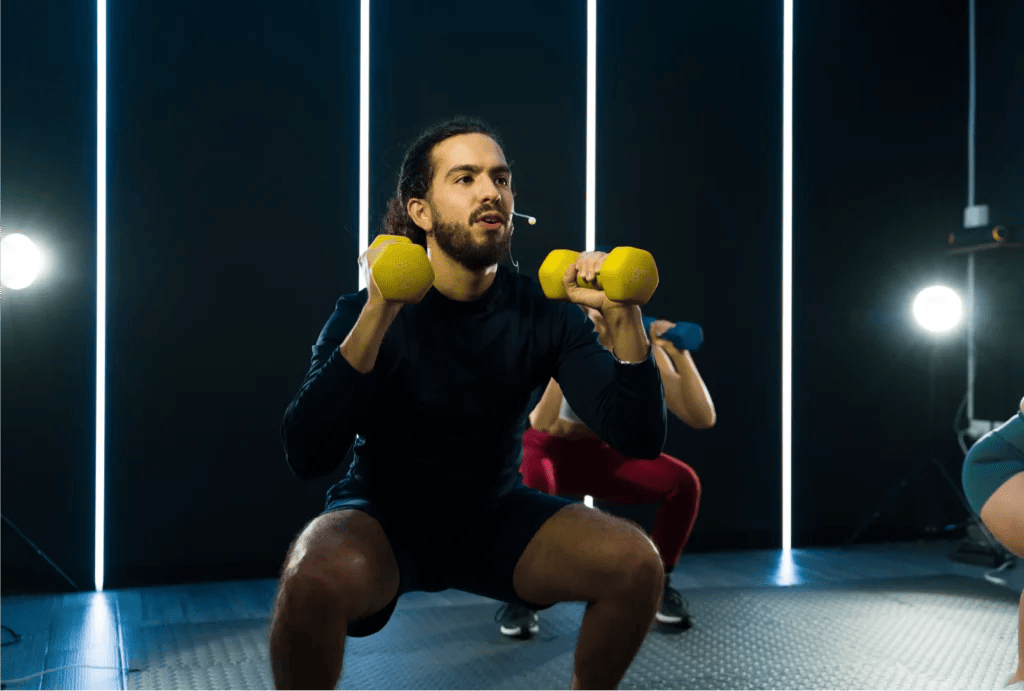 A man leading a workout class squatting holding two yellow dumbells