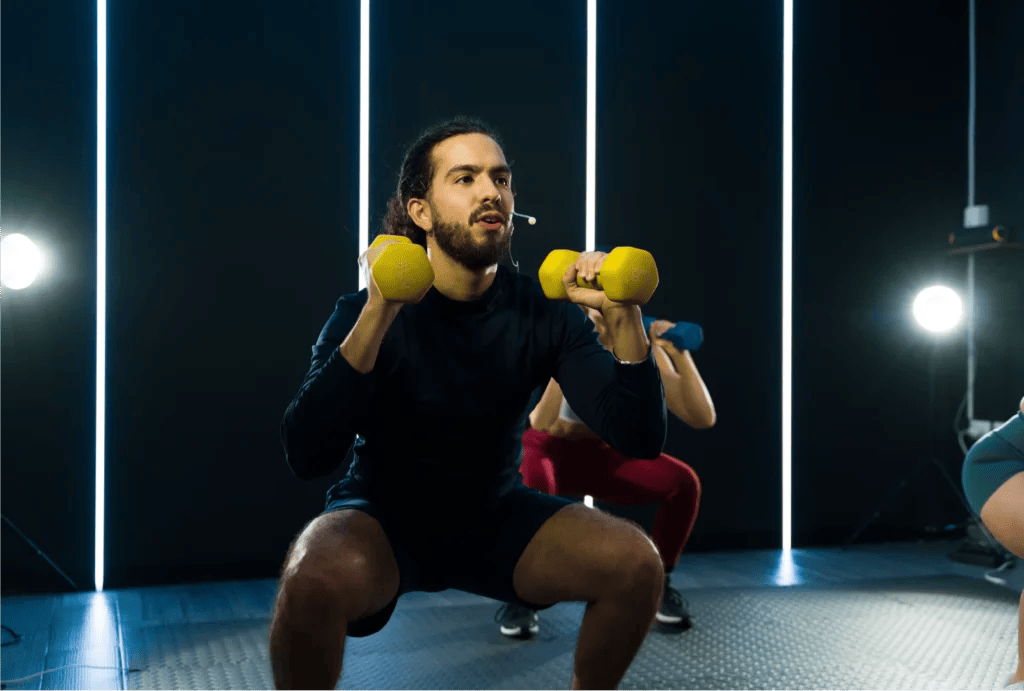 A man leading a workout squatting holding two yellow dumbells