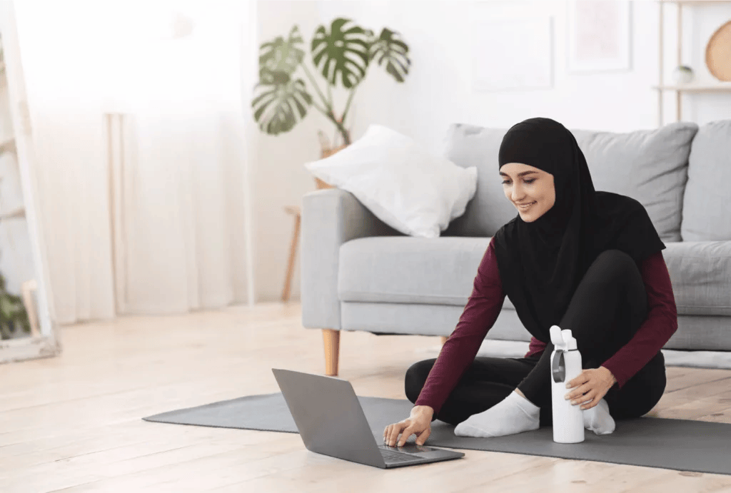 A woman wearing a hijab sitting on a yoga mat preparing for a workout session on her laptop