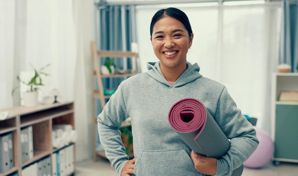 A woman in a sweatshirt smiles with a rolled up yoga mat in an indoor studio.