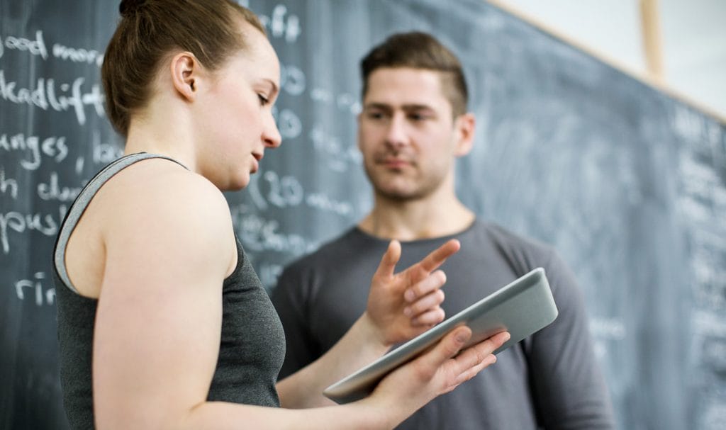 A woman and a man stand in front of a blackboard and talk while the woman looks at a tablet in her hand.