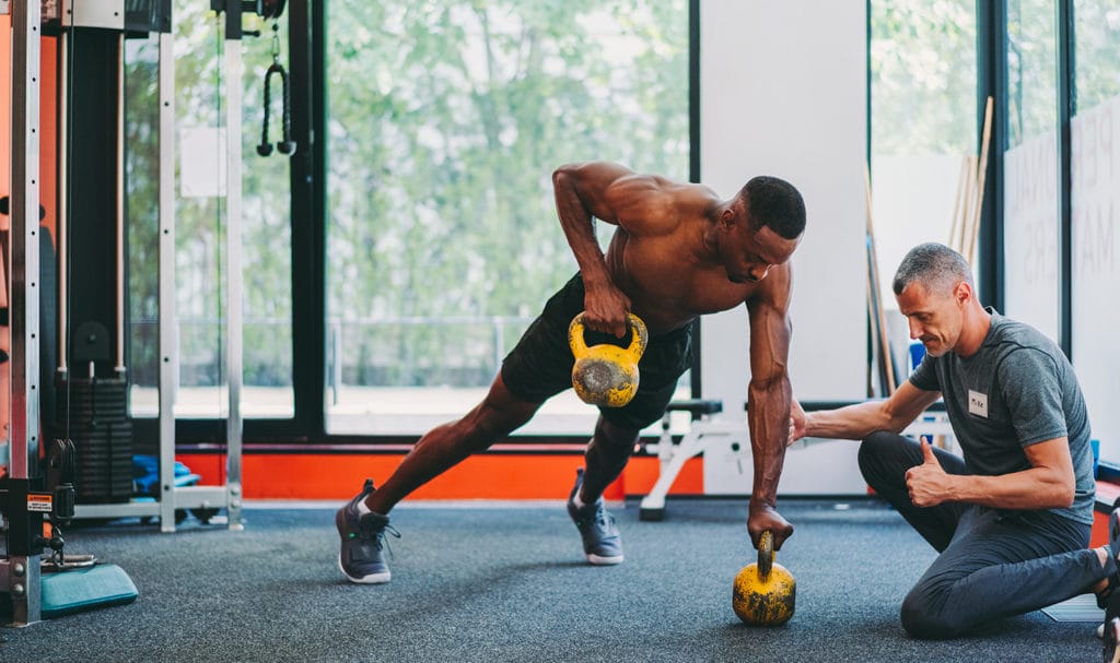 A male personal trainer is kneeling down on a workout mat as he helps his male client in a lunge position lift weights and improve his performance.