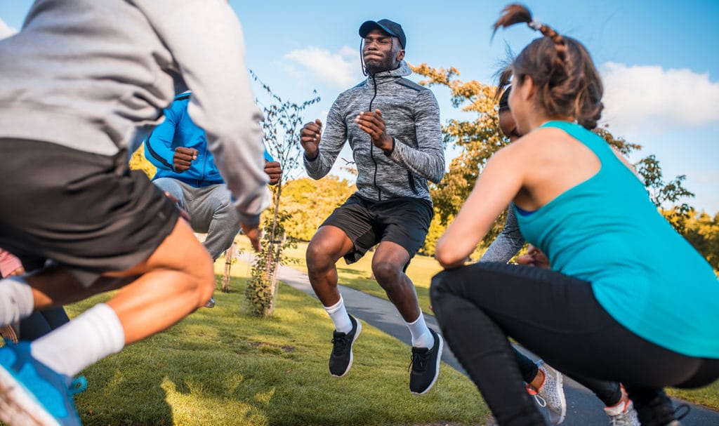 A personal trainer and his clients are all mid-jump as they perform HIIT exercises together outside.