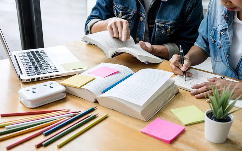 A prepared tutor sits at a desk next to a student, helping a them study for exams.