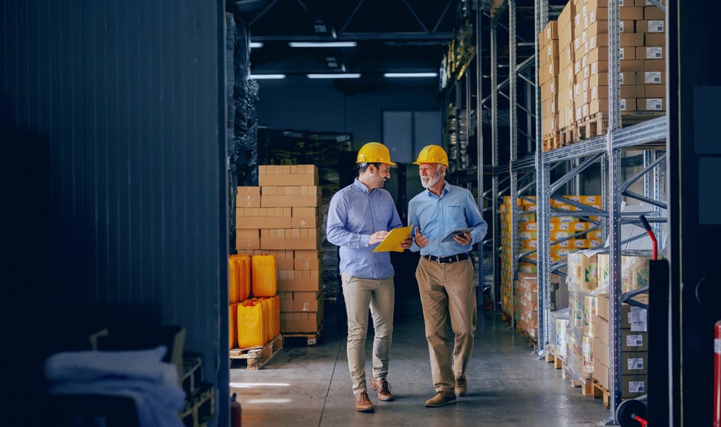 Two men are walking side by side discussing general and product liability insurance. They are wearing dress pants with blue dress shirts with yellow hard hats as they walk amongst shelves of boxed products in an industrial warehouse as they discuss product liability insurance.