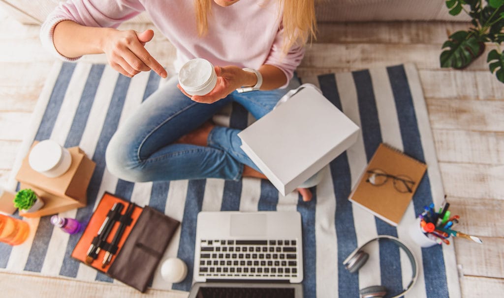 A woman sits with her legs crossed on a striped mat on the floor as she studies her different beauty products next to her laptop so she can get an insurance quote.