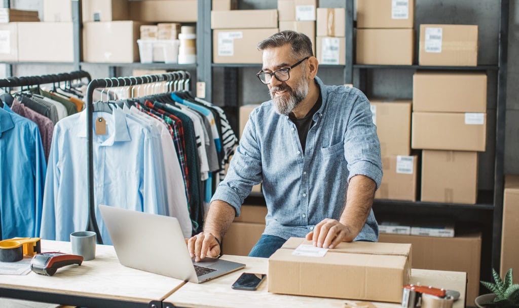 An older man works on his laptop to buy product liability insurance as he works in his home office surrounded by boxes and racks of products.