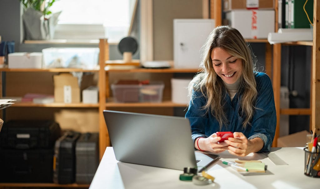 A young woman is chatting on her phone next to her laptop and paper work to an insurance agent about getting product liability insurance.