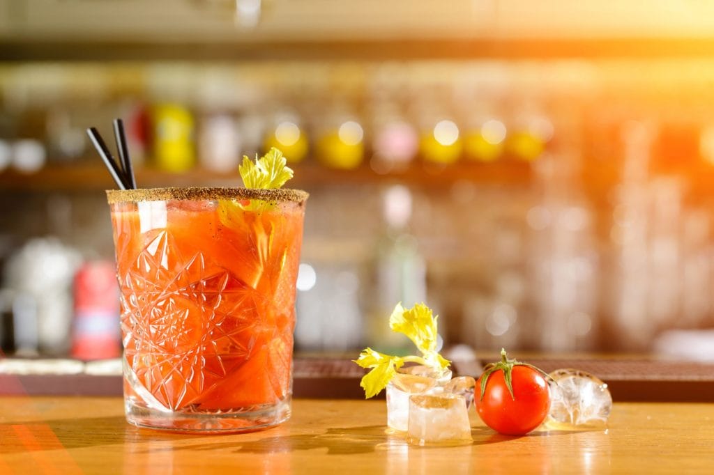 Close-up styled shot of mini bloody mary next to ice cubes, a cherry tomato, and celery leaves on a bar counter top.