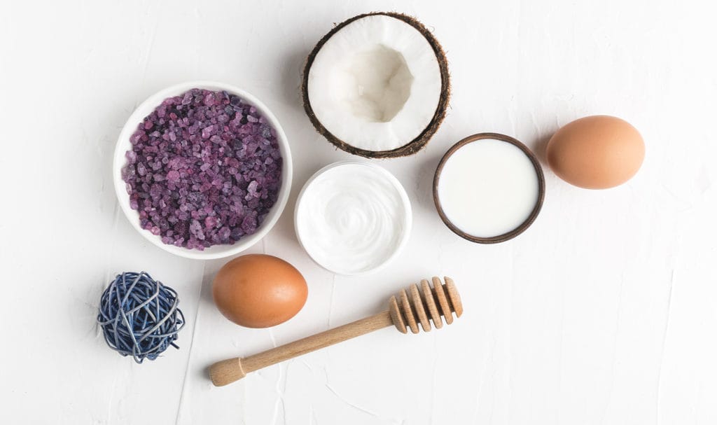 A coconut, eggs, creams, and flower petals lay on a white background to show the ingredients in a product.