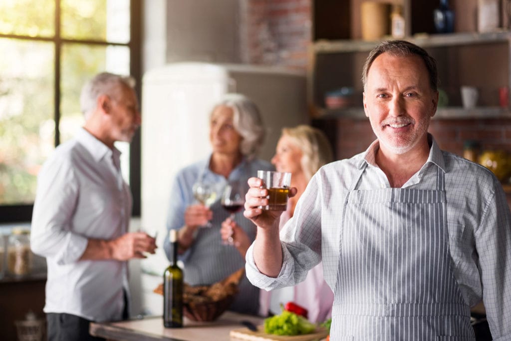 A man in an apron raises a cocktail while a group of friends talk and enjoy beverages in the background.