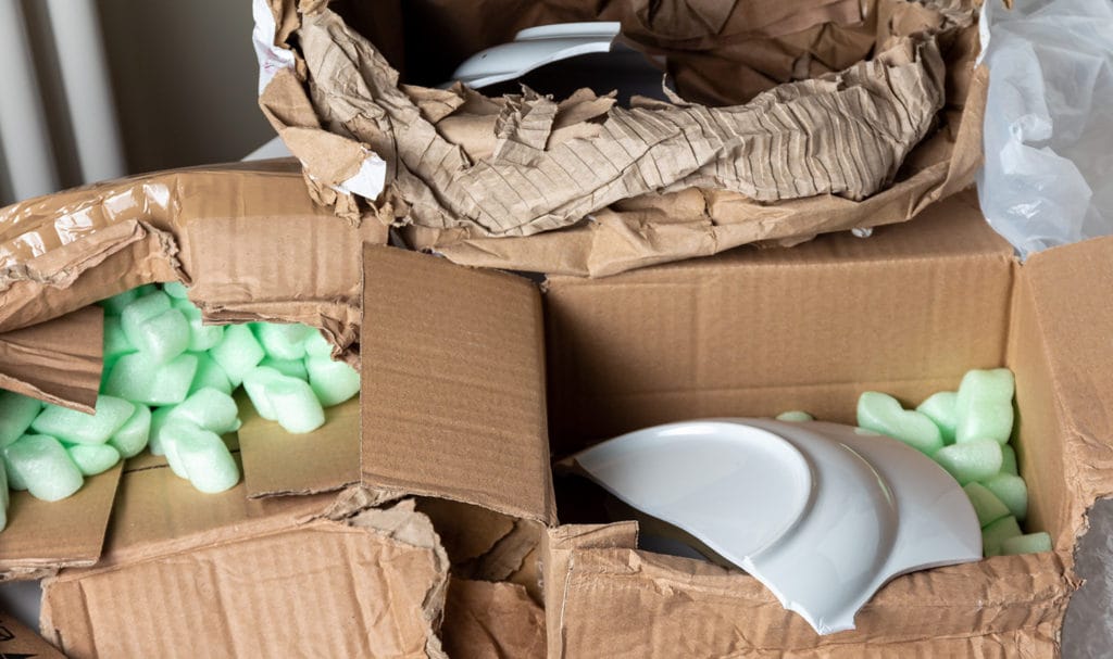 A stack of broken brown mailing boxes with broken white porcelain plates inside.