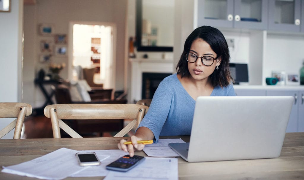 A woman is in her home sitting in her kitchen at the table with a laptop, paperwork, and a calculator as she calculates her risks.