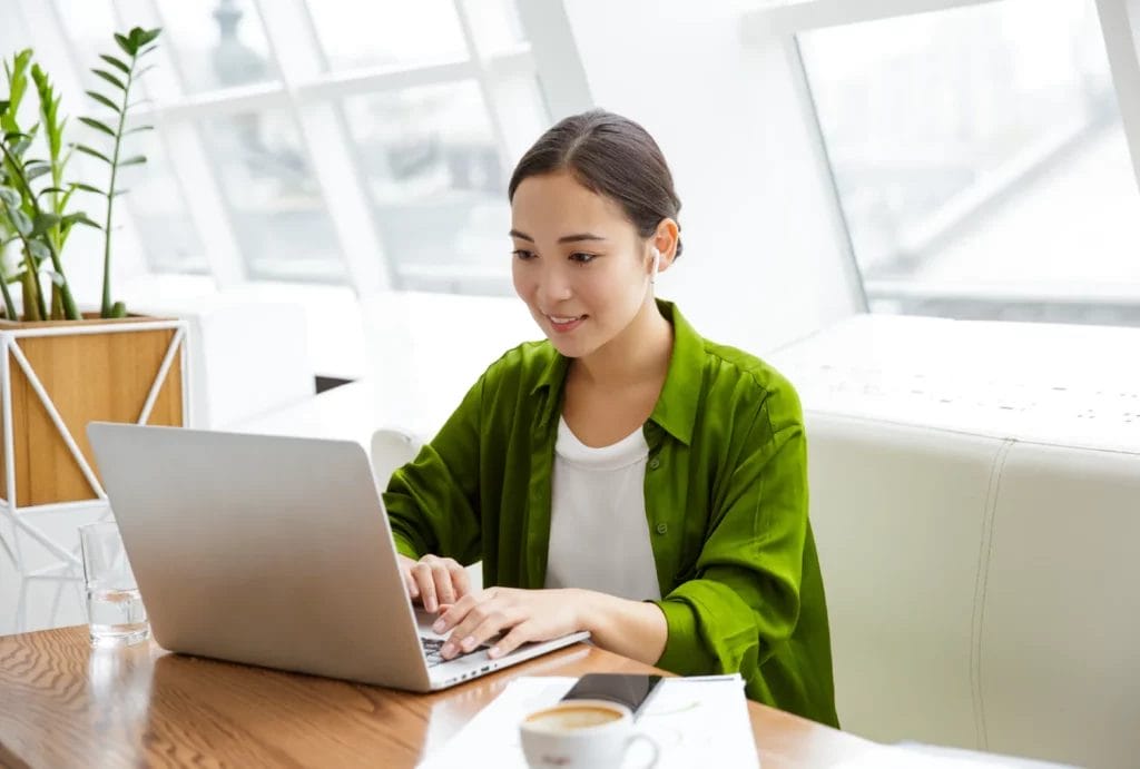 A woman wearing a green shirt smiles and looks at her smartphone while sitting at a wooden table with her laptop, coffee, and business papers in front of her.