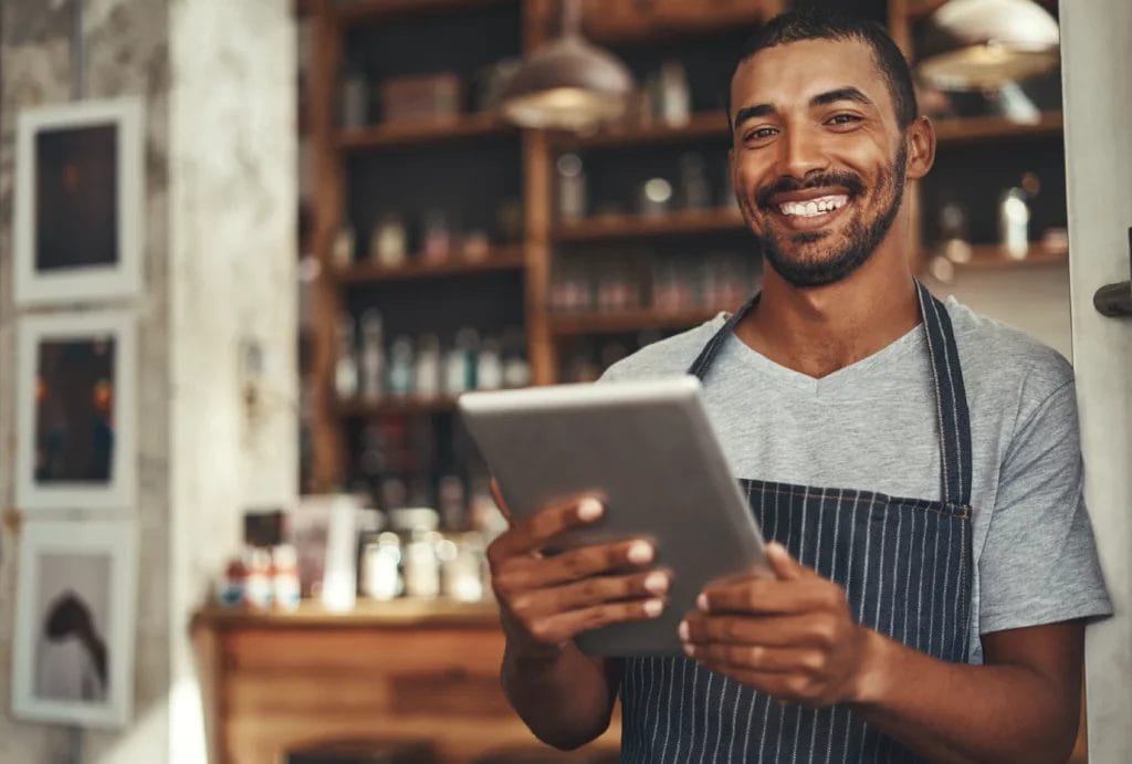 A small business owner wearing a gray shirt and denim apron holds a business tablet and smiles while leaning against a doorframe.