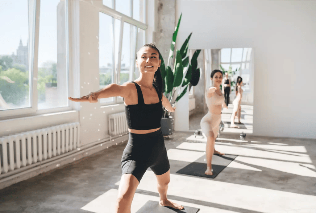 A yoga teacher wearing a black athleticwear seat holds warrior three pose while teaching class in a bright studio with large glass windows in the background.