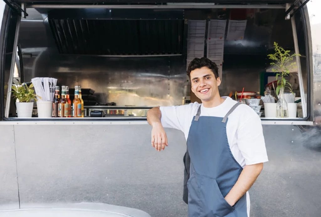 A food truck owner wearing a white shirt and blue apron smiles and leans against his food truck window.